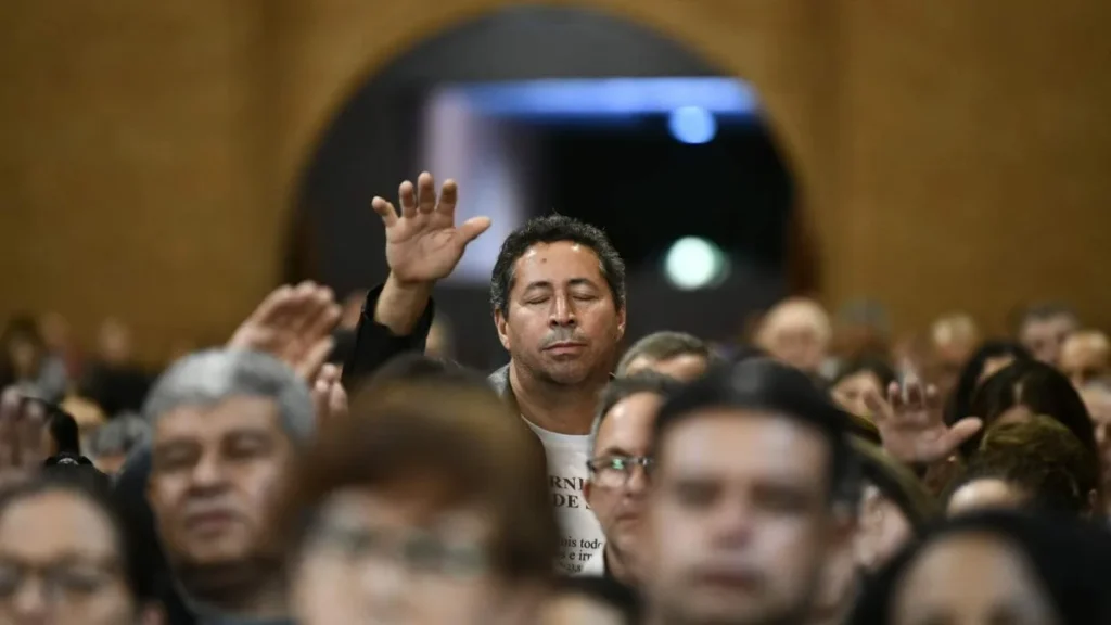Imagem devoto reza de olhos fechados e mao estendida no meio do povo no 7 dia de novena solene do artigo Adorar ou venerar: qual a diferença para os católicos?