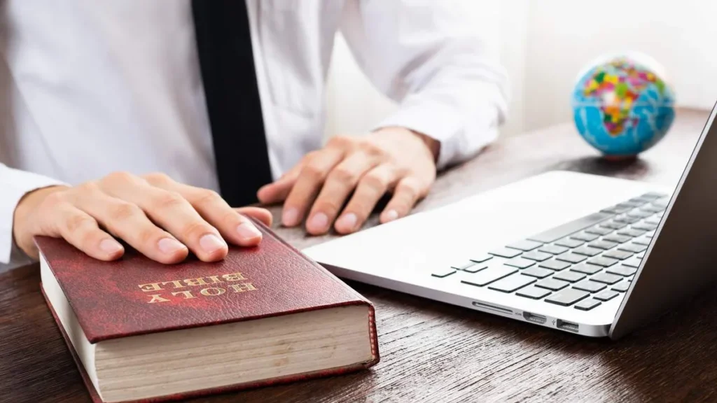 Imagem maos de homem sobre a Biblia que esta em cima d euma mesa e um notebook ao lado do artigo A evangelização na era digital