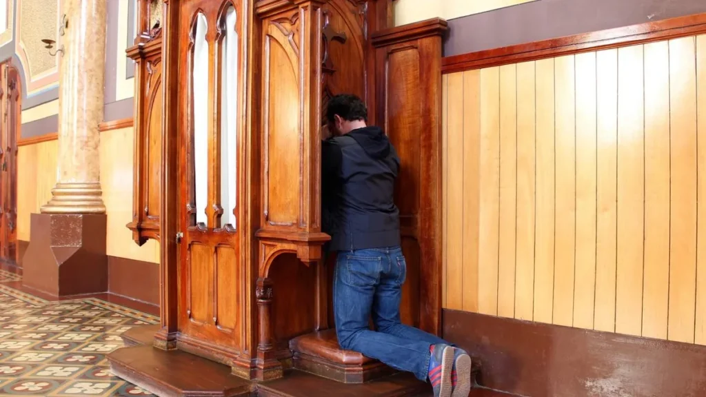 Imagem homem ajoelhado numa igreja se confessando com um padre na cabide de confissao do artigo Por que se confessar? Entenda o sacramento da reconciliação