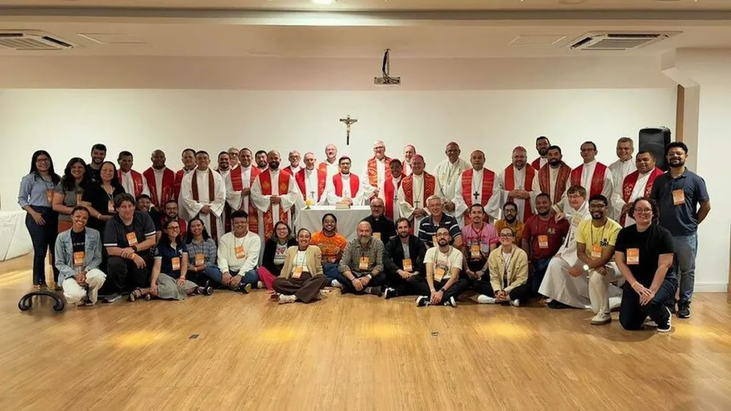 Imagem Grupo de pessoas a frente de uma parede branca posicionados para a foto. Esse grupo representa a Comissao para a Juventude do artigo CNBB dá passo importante para a evangelização da Juventude