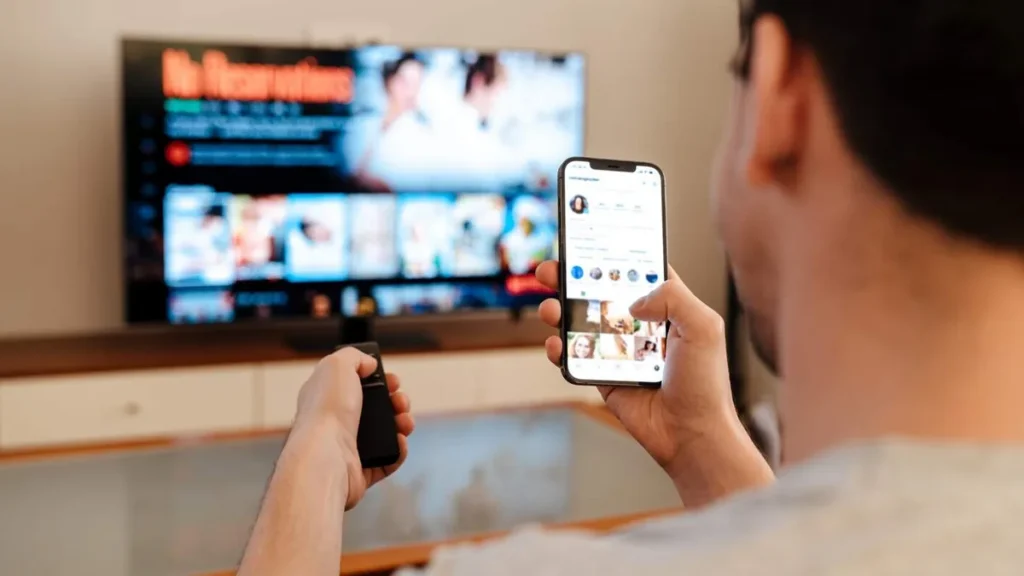 Imagem homem com celular na mao mexendo na tv com o controle remoto do artigo Como simplificar a vida em tempos de consumo